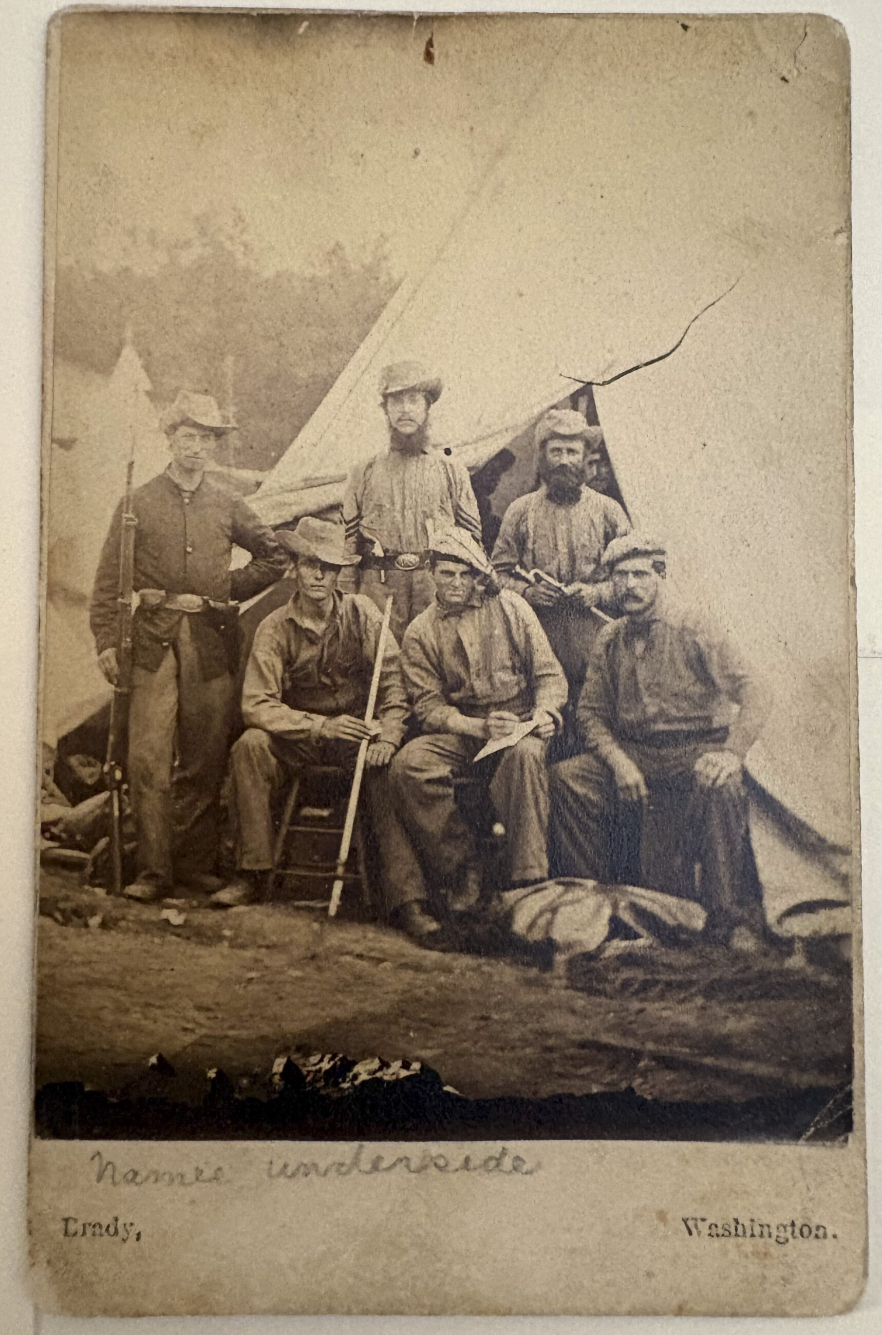 A group of soldiers in mid 1800s dress -- hats and simple clothing -- in front of a tent.