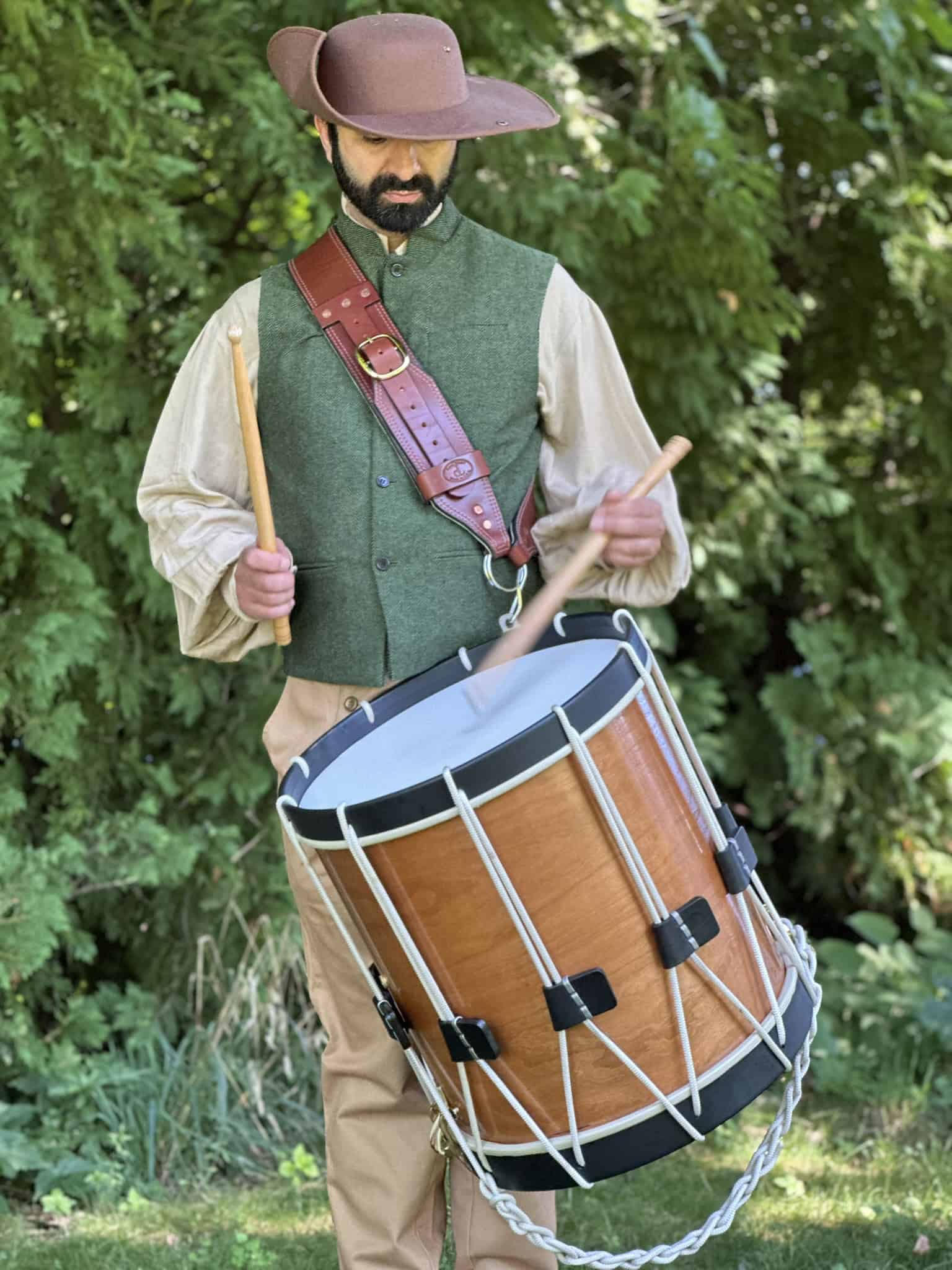 Photo of a man in historic costume, including a brown hat and green vest, playing a large rope drum worn with a leather strap across his chest