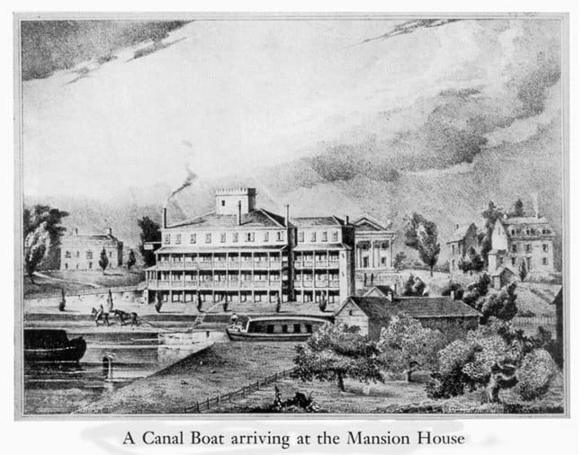 A detailed black-and-white historical engraving depicts a long, flat-bottomed passenger canal boat arriving at the Mansion House, a grand four-story hotel dominating the center of the scene. The hotel features extensive multi-level open-air verandas running across its facade, smoking chimneys, a small rooftop cupola, and a classical Greek Revival wing with columns on the right. In the foreground, the calm waters of a man-made canal bend wrap around the property, where the covered passenger boat is moored at the landing basin, and a second barge sits near the left bank. On the left side of the waterway, a horse-drawn carriage travels down a dirt path, while the right foreground is filled with lush trees and small wooden outbuildings.