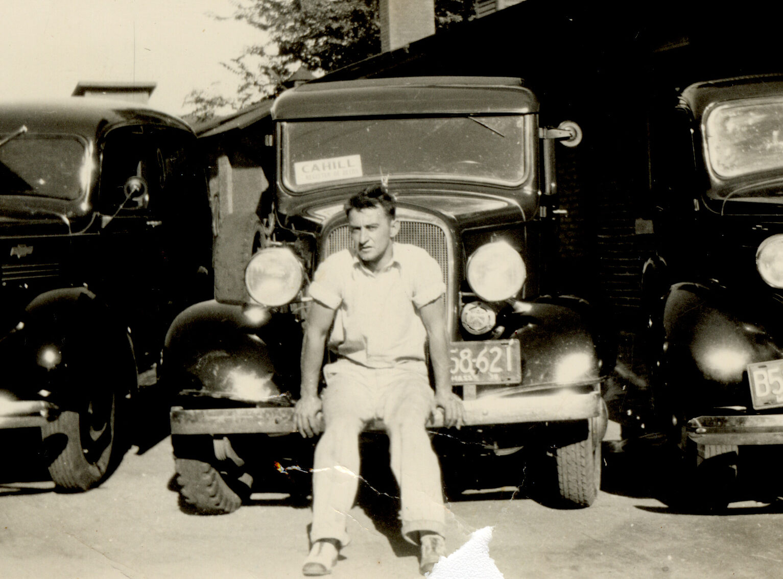 Black and white photograph in "The Belt" neighborhood. Young man leaning on a car, two other cars next to him.