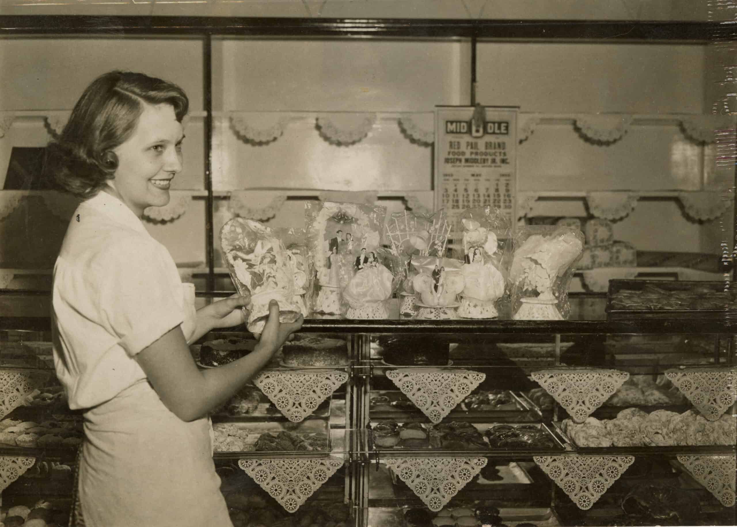 black-and-white photograph of a young woman working inside a bakery shop, surrounded by baked goods and elaborate wedding cake toppers. Standing on the left in profile, the smiling woman has styled, shoulder-length wavy hair and wears a light-colored, short-sleeved blouse paired with a matching apron. She holds a decorative, cellophane-wrapped cake topper in her hands, carefully placing or arranging it on top of a glass display counter. A row of several other wrapped wedding toppers, featuring miniature bride-and-groom figurines and floral arches, is lined up along the counter surface. Below, the glass display cases are packed with rows of pastries, cookies, and baked treats, each shelf neatly lined with white, triangular, lace-patterned doilies hanging over the edges. In the background, a white wall features empty display shelves with scalloped paper lining. Hanging prominently on the back wall is a calendar featuring a bold logo at the top that reads "MIDDLE / RED PAIL BRAND / FOOD PRODUCTS / JOSEPH MIDDLEBY JR. INC."