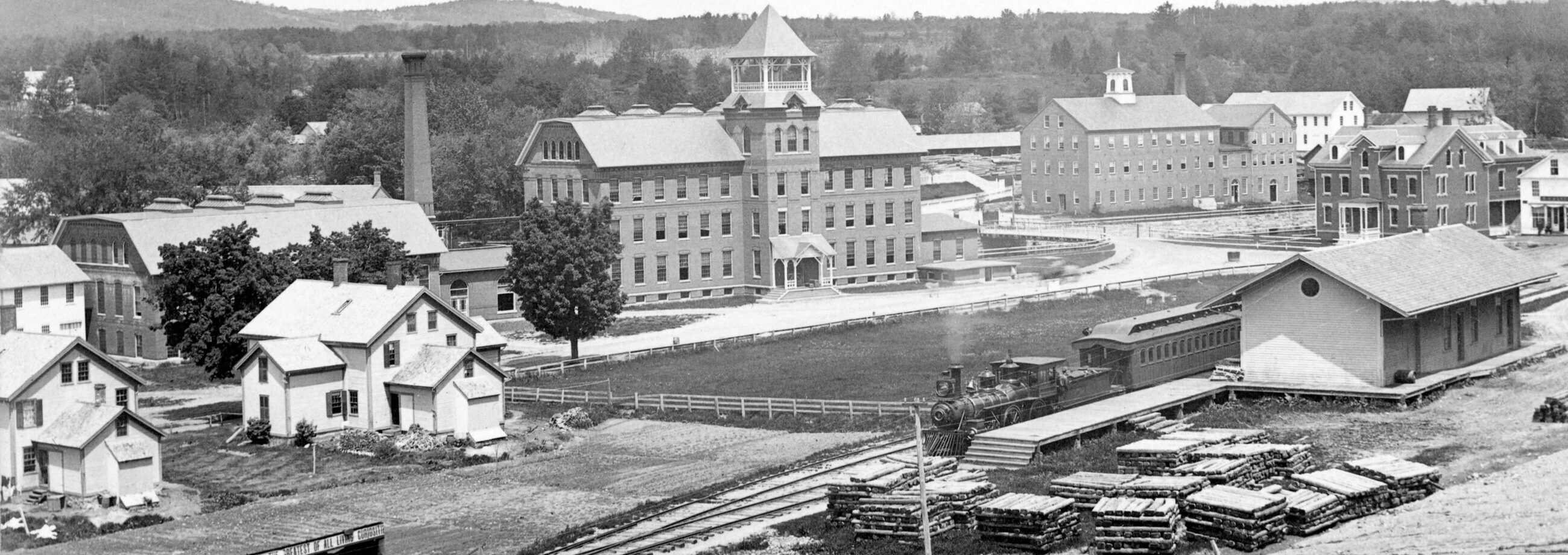 Black-and-white photograph of a 19th-century industrial complex with rolling, tree-covered hills in the background. In the foreground, a steam train moves along a dirt railway bed, passing a long wooden platform loaded with numerous organized stacks of lumber. To the right of the tracks stands a simple, light-colored wooden train depot with a gabled roof and a dark circular window on its side profile.The center of the image features a massive, three-story brick mill building with a prominent central tower topped by an open-air belfry and a pointed roof. A large, dark smokestack rises high into the air behind a long, multi-section factory building situated to the left of the main mill. Scattered across the rolling dirt and grass landscape are various other structures, including several multi-story stone or brick industrial buildings in the upper right, and a few small, two-story white residential homes with dark gabled roofs spread throughout the lower left.