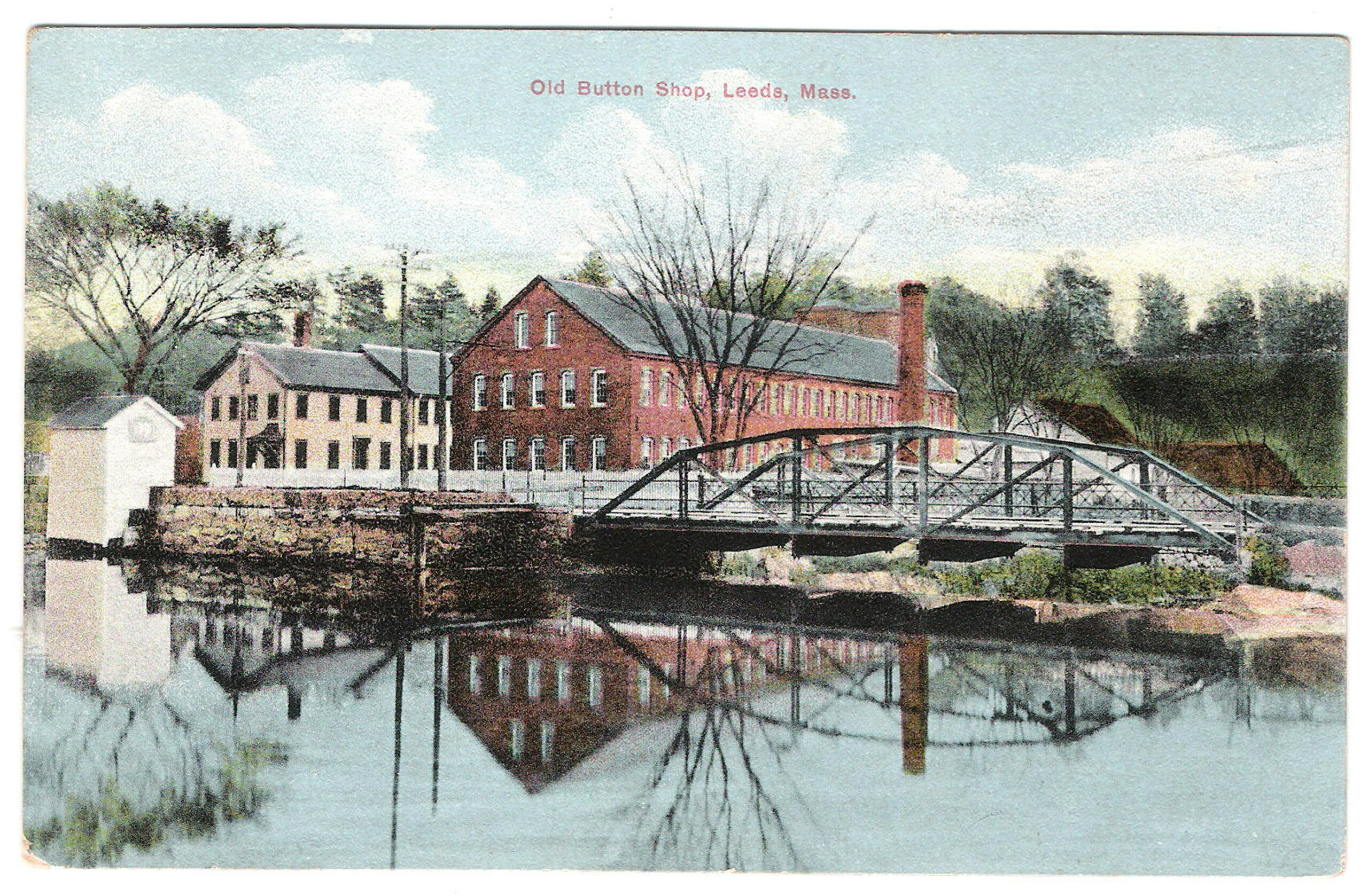 A color postcard depicts a scenic view of the "Old Button Shop" situated along a calm body of water. In the center, a prominent two-story red brick industrial building features rows of small, rectangular windows, with a smaller white-painted building attached to its left side. To the far left, a small, white wooden structure sits directly on the stone-walled bank of the river, its reflection clearly mirrored in the still water below. A gray metal truss bridge spans the river from the right side, leading toward the brick mill complex. The foreground consists of glassy, dark water perfectly reflecting the brick buildings, the bridge, and the surrounding bare trees. Lush green trees and a lightly forested hillside rise in the background under a pale blue sky filled with soft, white clouds. Red text printed centered at the top reads, "Old Button Shop, Leeds, Mass."