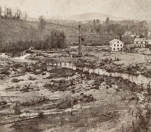 Photograph documents the extensive devastation following the 1874 Mill River Flood in Williamsburg, Massachusetts. The foreground and middle ground consist of a wide, barren valley of eroded mud and sand heavily littered with uprooted tree roots, scattered branches, and debris. Near the center of the image, a lone, tall brick factory smokestack stands isolated amidst the ruins where an industrial mill once stood. To its right, a white two-story wooden house with a small rooftop cupola remains upright, situated next to another residential building farther back, both surrounded by the mud plain. A shallow, narrow stream of the Mill River snakes through the center of the washed-out landscape, spanned by the precarious remains of a broken wooden footbridge. In the background, a steep, partially cleared hillside rises on the left, leading to distant rolling hills and a dense tree line