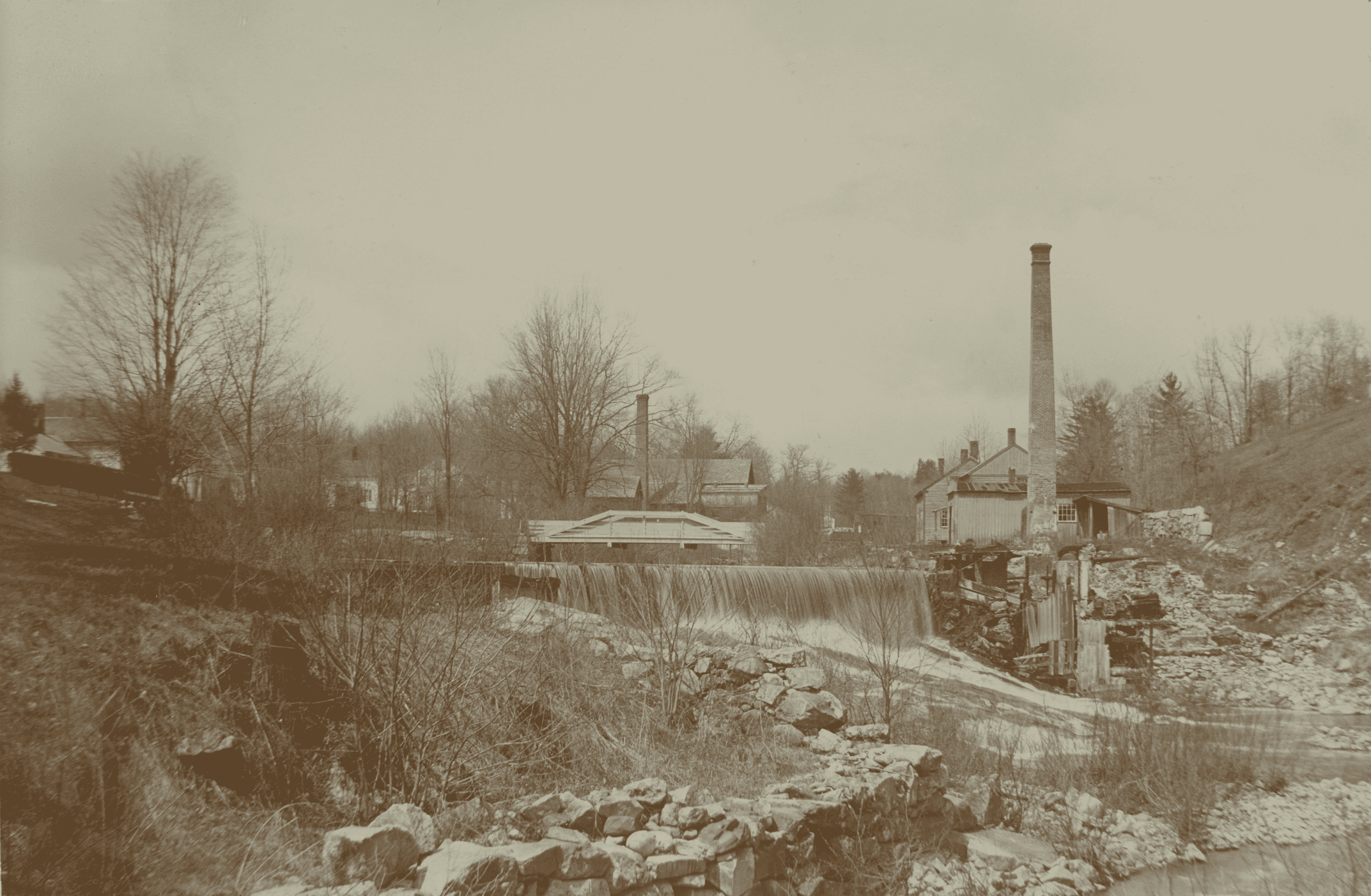Black-and-white image of a rural industrial scene. In the midground, a wide dam spans a river, creating a smooth cascade of water that flows toward the right. To the right of the dam, a group of old, wooden mill buildings stands along the rocky bank, featuring a prominent, tall brick smokestack rising high into the pale sky. Another smaller chimney peaks out behind the central trees. Surrounding the mill and the river are bare, winter trees and a gentle slope on the far right. The foreground is composed of a rough, crumbling stone wall and dry brush overlooking the river. In the distant left background, a few small houses are nestled among sparse trees.