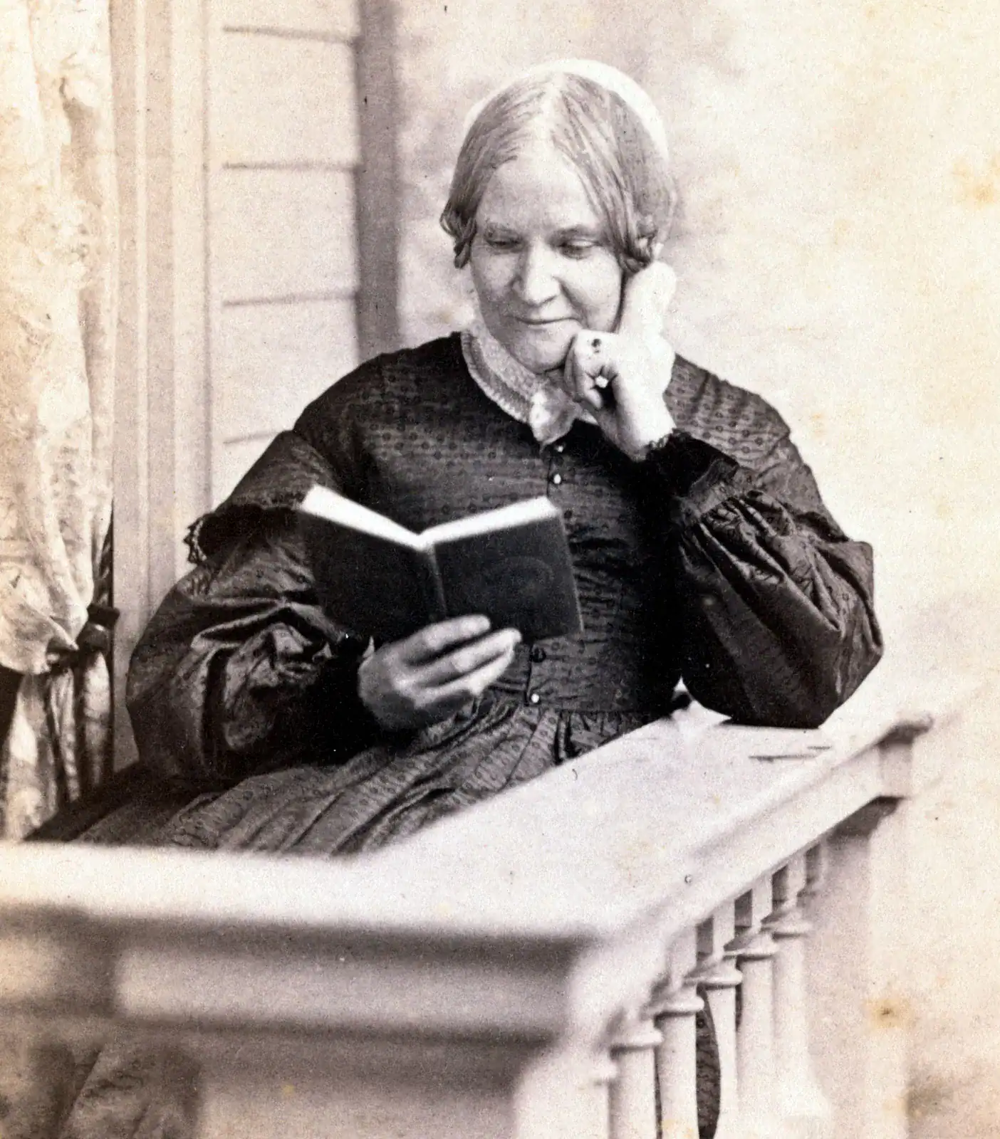 Black-and-white photograph of Lydia Maria Child posing while reading a book. She rests her head on her hand and smiles down at her book. She wears a black dress with large puffy sleeves. She sits outside in a small porch-like or balcony area.