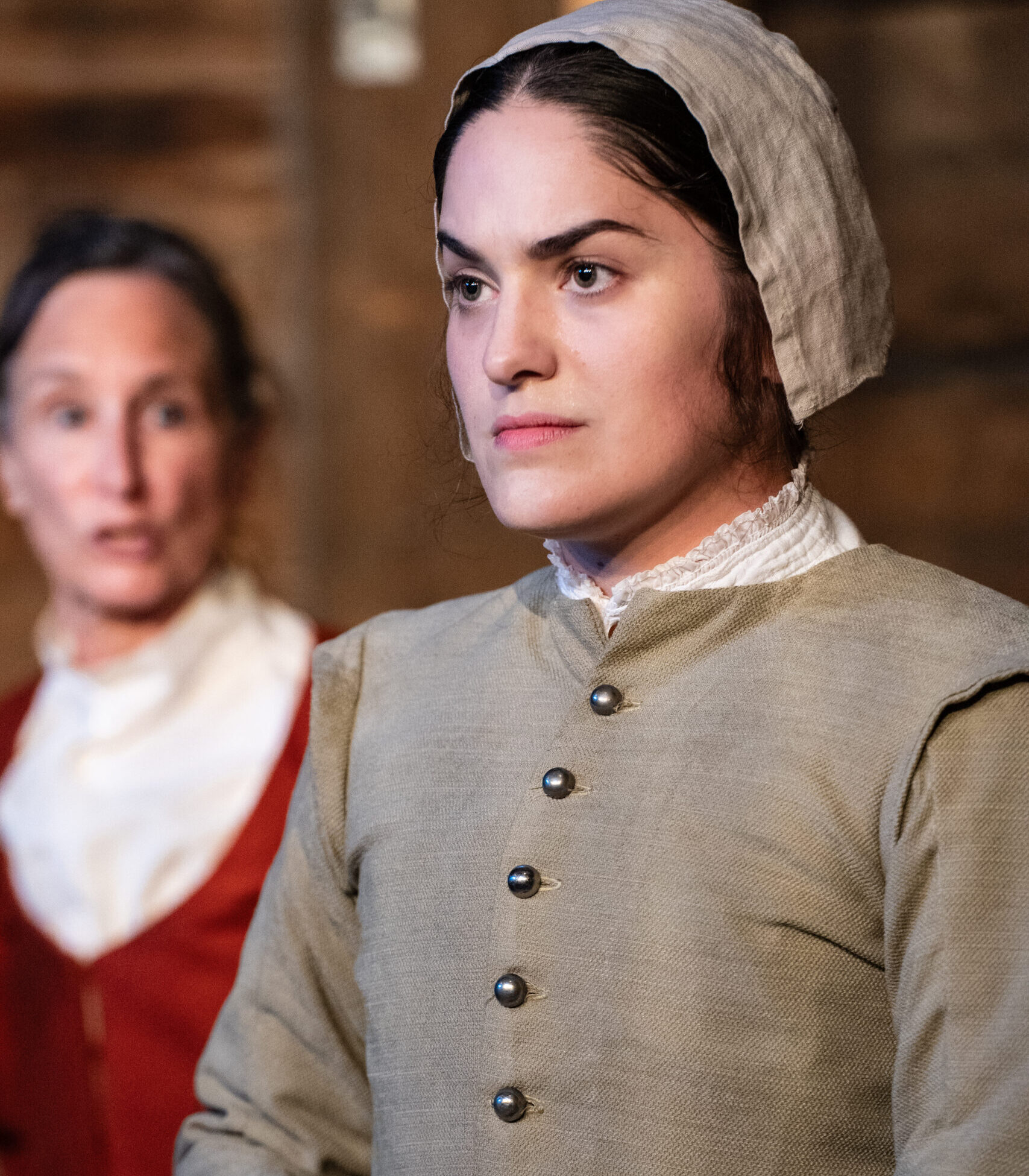 Photograph focuses on Sarah Lyman Bridgman (Linda Tardif) looking sternly into the distance. She has on a light brown buttoned up dress and a white head covering. In the distance, Mary Bliss Parsons (Christine Stevens) can be seen glaring at Bridgman appearing to be talking angrily or fearfully. She wears a red and white dress.