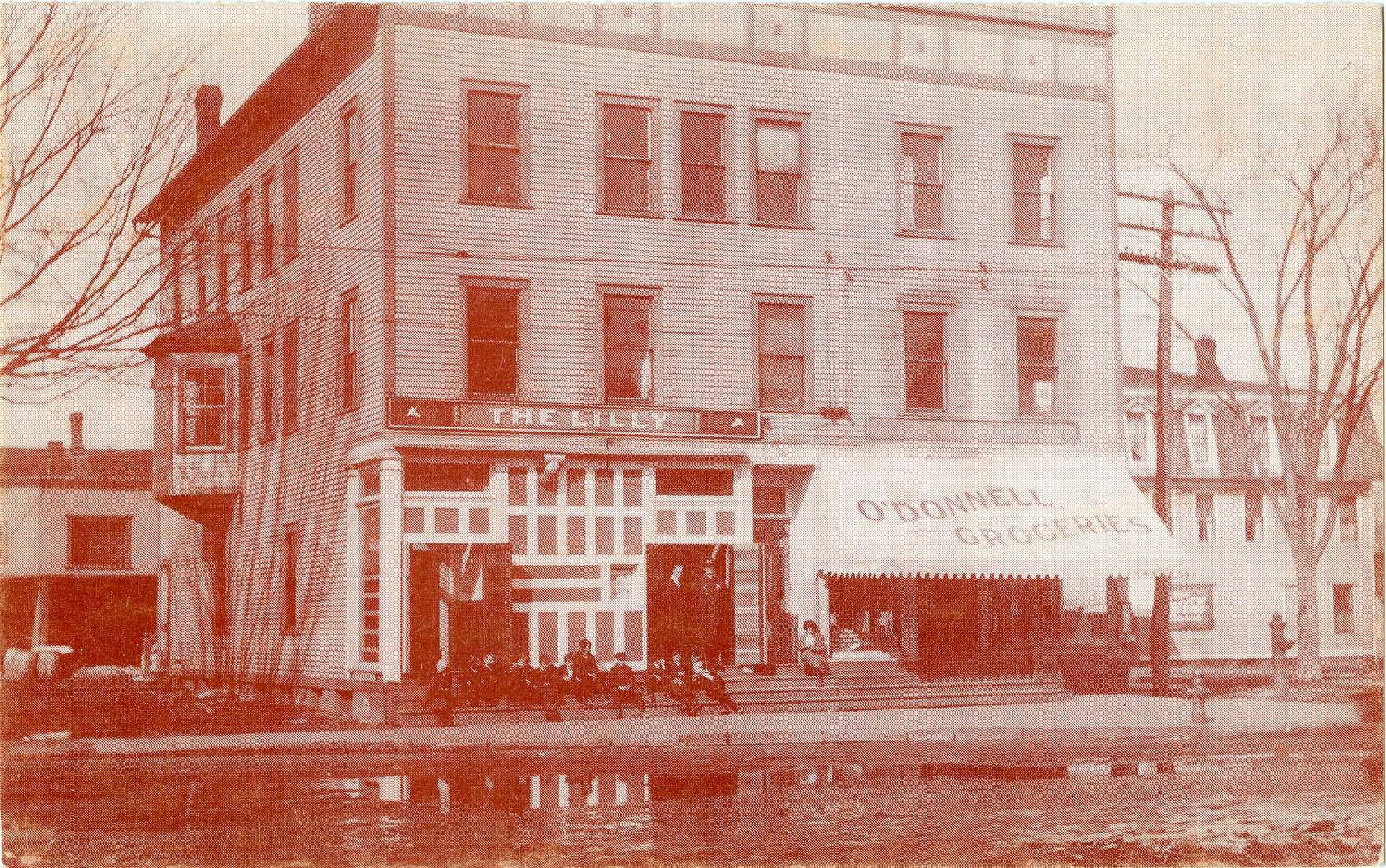A black-and-white photograph of Maine Block. Storefronts are in view with signs reading: "THE LILLY" and "O'DONNELL GROCERIES." Young boys are sitting on front steps.