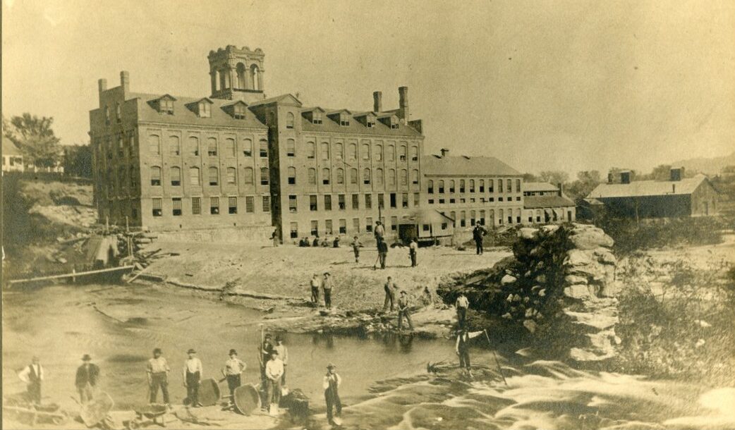 sepia toned photograph of a factory building on the edge of a river with a damn across. Many men are standing on and around the dam, some with tools.
