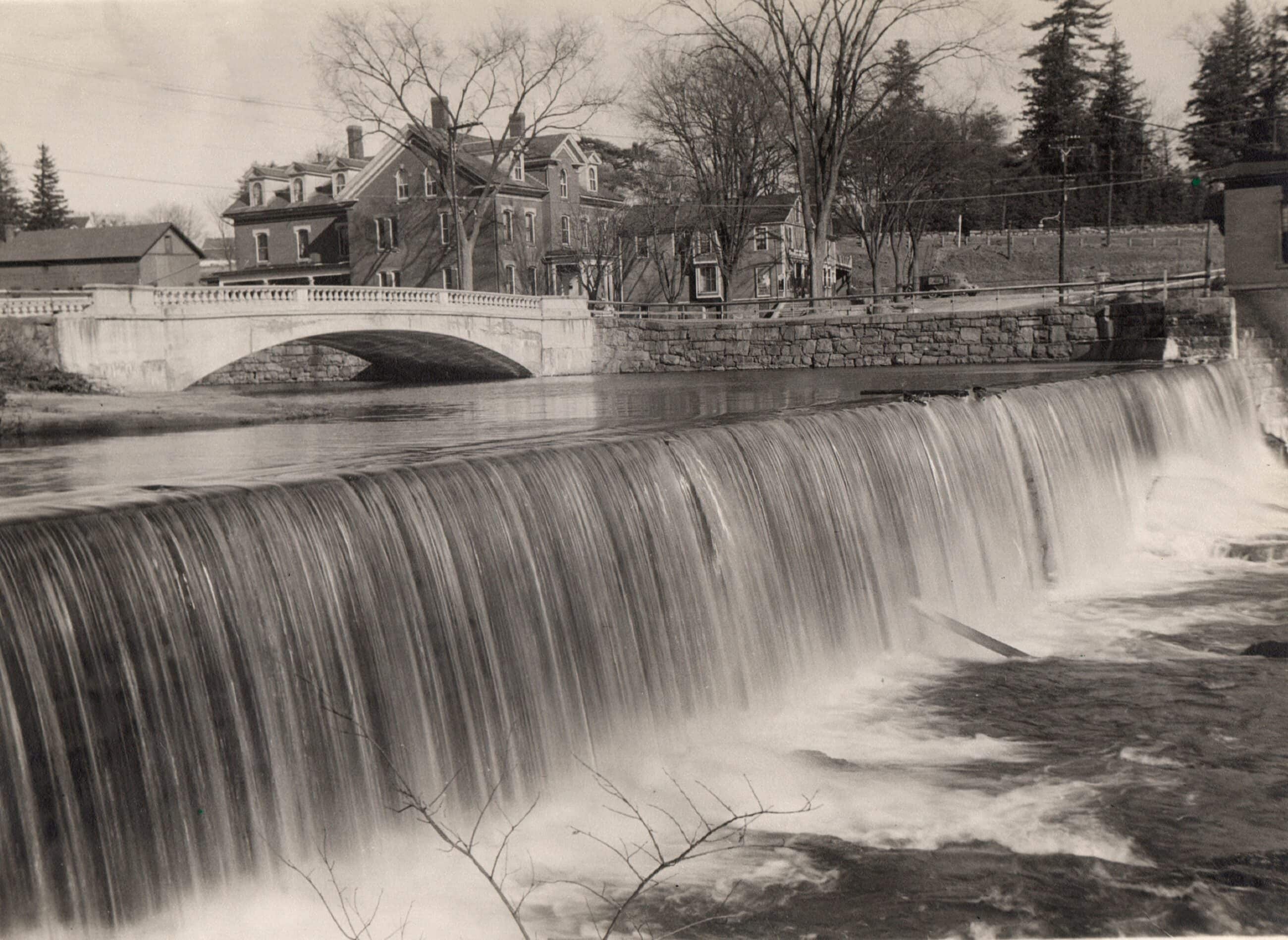 Black and white photograph shows a view of a river dam. In the foreground, a large, stone dam stretches horizontally across the river, creating a wide waterfall where water cascades smoothly over the edge into a turbulent, foaming pool below. A few bare tree branches peek into the bottom center of the frame.
Behind the dam, the river water remains calm and flat. Further back on the left, an arched stone bridge with an elegant balustrade spans across the river channel. On the far side of the river, a stone retaining wall lines the bank. Nestled behind the wall and the bridge are several multi-story historic buildings with pitched roofs, including a prominent brick house with dormer windows. Bare trees are scattered throughout the town landscape, and a line of evergreen trees stands on a distant hill