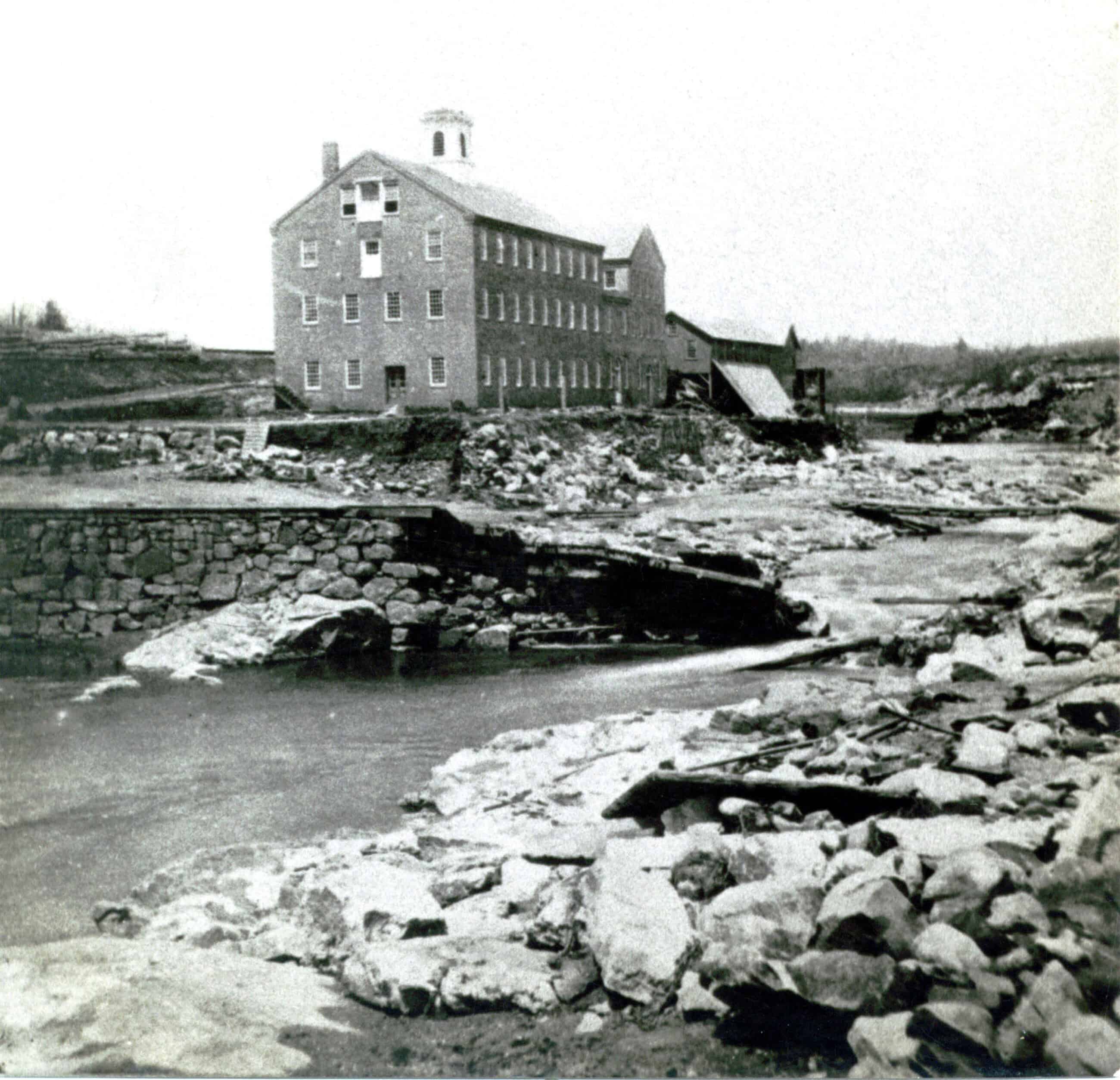 A black-and-white photograph captures a multi-story brick mill building situated along a wide, rocky riverbed. The large, rectangular building features three main floors and an attic level with rows of uniform multi-pane windows, and a small, white cupola sits atop its gabled roof. In the foreground, the river flows amidst heavy devastation, with its banks severely eroded and scattered with large boulders, loose rocks, and broken timber. To the left, a partially collapsed stone retaining wall or bridge structure sits broken near the water's edge, while the background reveals bare hillsides and a few smaller damaged wooden structures connected to the main mill complex.