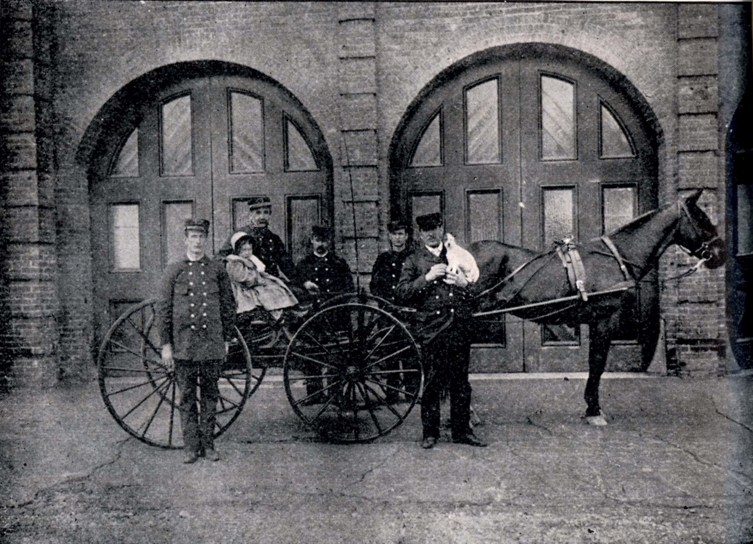 Black-and-white photograph of five men in button down black suits next to a horse and carriage with a small child sitting in it. They are infront of the carriage entrance of the fire station.
