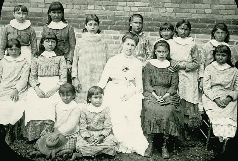 Class portrait of Native American girls appearing to be around preteen age standing and sitting in three rows. Their expressions are all serious. Most of the girls wear long-sleeved, loose-fitting dresses or smock-like garments made of patterned or checkered fabric. In the front center is their teacher, a white woman dressed in white, slightly smiling at the camera.