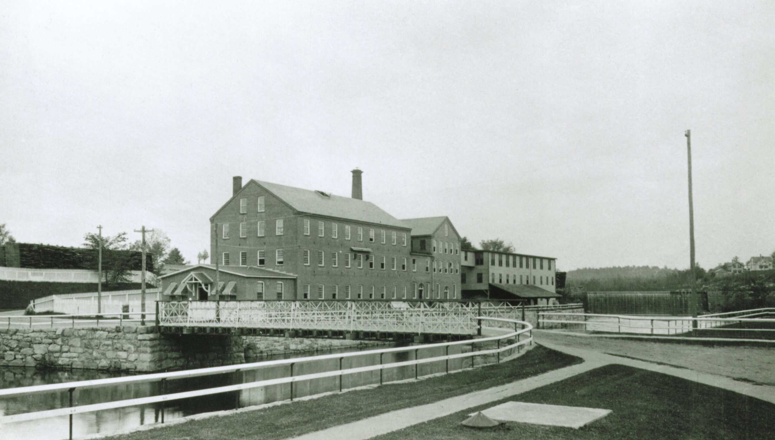 Black and white photograph captures an industrial mill complex alongside a river and canal system.
In the center foreground, a stone-lined canal flows from left to right, bordered by a dark path and a light-colored safety railing that curves along the water's edge. A low, ornate white lattice truss bridge spans across the canal, connecting the foreground pathway to the main mill grounds.
Dominating the center of the frame is a large, multi-story brick mill building with a gabled roof and a prominent central chimney stack. Rows of multi-paned rectangular windows line its facade, and a smaller, single-story entrance vestibule with an awning extends from its left side. Connected to the rear of this main structure is another long, multi-story industrial building that stretches toward the right.
In the background on the far right, a low dam controls the river's flow, while rolling hills and sparse residential buildings sit on the distant tree-lined horizon. Utility poles stand on both sides of the canal.