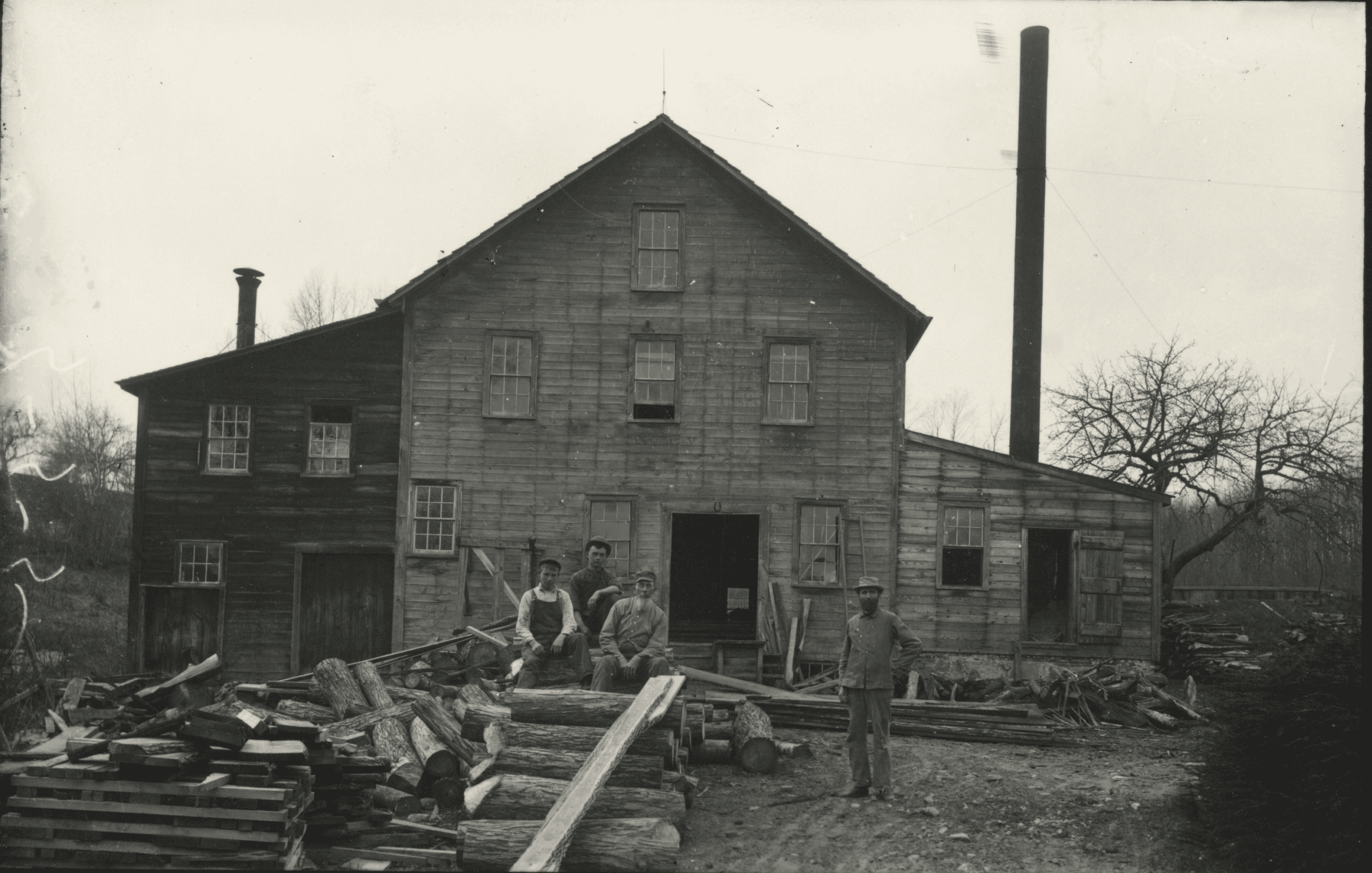 Black-and-white image of a multi-story wooden mill building with several workers posing outside amidst scattered lumber. The central structure is a weathered, rustic three-story building constructed from horizontal wooden planks, featuring a prominent gabled roofline and multiple small, multi-pane windows distributed across its facade. On the left side, a darker wooden addition with an uneven roofline blends into the main structure, while a single-story extension projects from the right, backed by a tall, slender black smokestack rising high into the sky. In the foreground, four men dressed in work attire are positioned near the building's main open doorway; three are seated together on a large pile of logs, while the fourth stands independently to their right. The ground in front of the mill is heavily cluttered with an active workspace's debris, including cut logs, long timber planks, and scattered wood scraps. In the background, bare, leafless trees frame the mill.