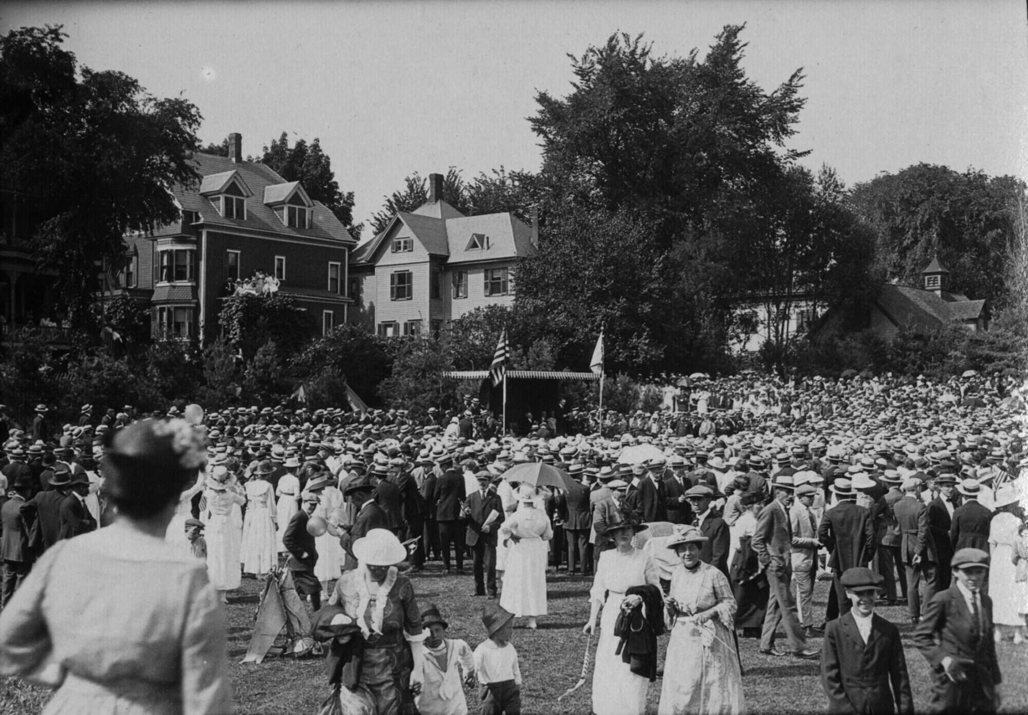 Black-and-white photograph showing a large crowd on a lawn at Smith College. In the distance, young people can be seen watching from the roof of their house.