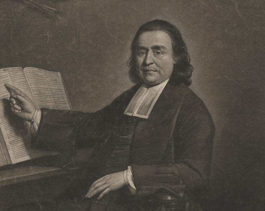 Portrait of man depicted from the waist up, seated and facing slightly to the right, looking directly at the viewer with a calm expression. He has shoulder-length, dark wavy hair and is dressed in dark ministerial robes with a traditional white clerical collar (preaching bands) at his neck. His right arm is extended to the left, with his index finger pointing distinctly toward an open, large religious book or Bible resting on a wooden stand. His left arm rests lower, with his hand placed near his lap against his clothing. The background is dimly lit and textured.
