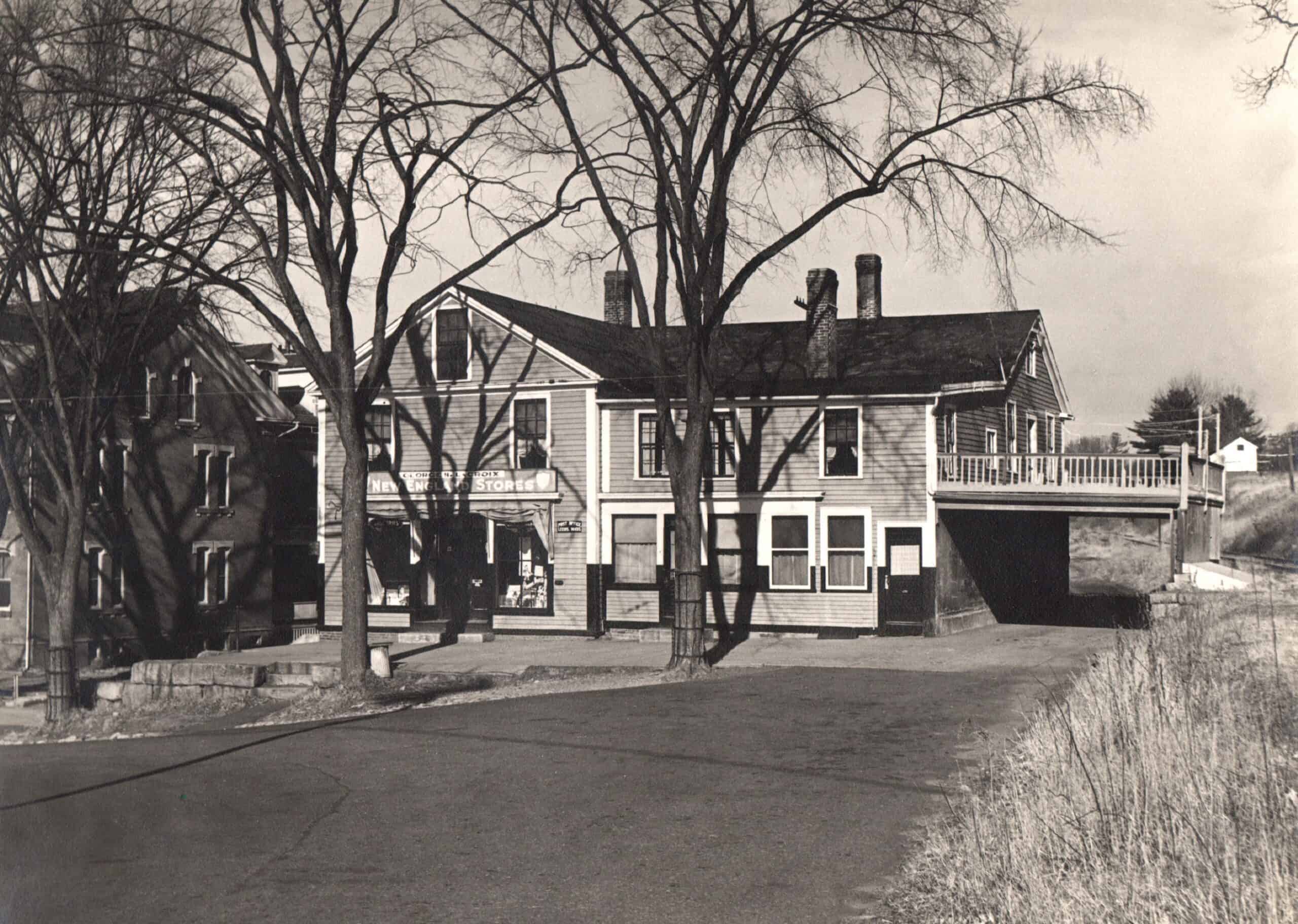 A black and white photograph shows an old, two-story wooden building facing a paved road, partially obscured by the bare branches of several tall, mature trees in the foreground. The building is divided into distinct sections; the left portion features a gabled roof with a storefront on the ground level, showing windows and a sign above them, while the right section has a flat roofline and includes a raised outdoor deck or balcony supported by posts on the far right end. To the left of the main structure, another dark, multi-story building is visible behind the trees, and the foreground consists of an empty asphalt driveway or parking area, with tall, dry grass lining the right side of the frame.
