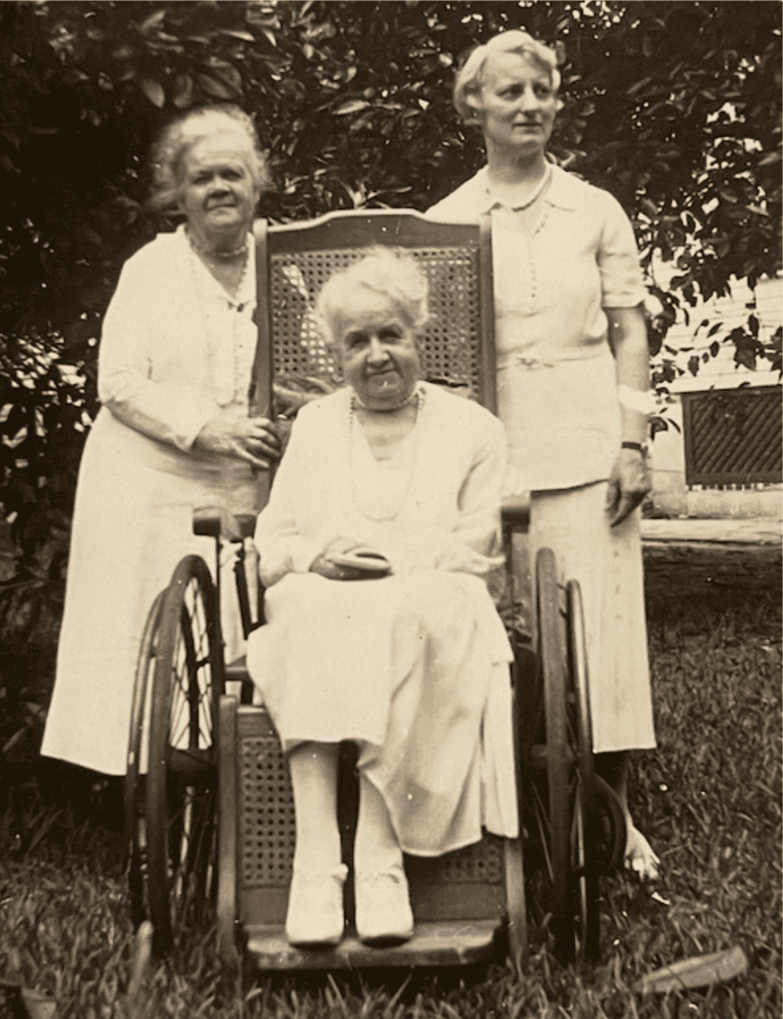 Black-and-white photograph of three woman outside infront of trees, two standing behind a woman sitting in a wheel chair. On the left stands a matured woman in a white dress with graying hair, holding onto the wheelchair infront of her. The woman in the wheelchair is matured with white hair. She is wearing a long white dress, and smiling toward the camera. The rightmost woman stands behind the wheel chair put looks off into the distance. She appears to be the youngest, around middle-age, and has light-colored hair. She wears a white dress coat and skirt.