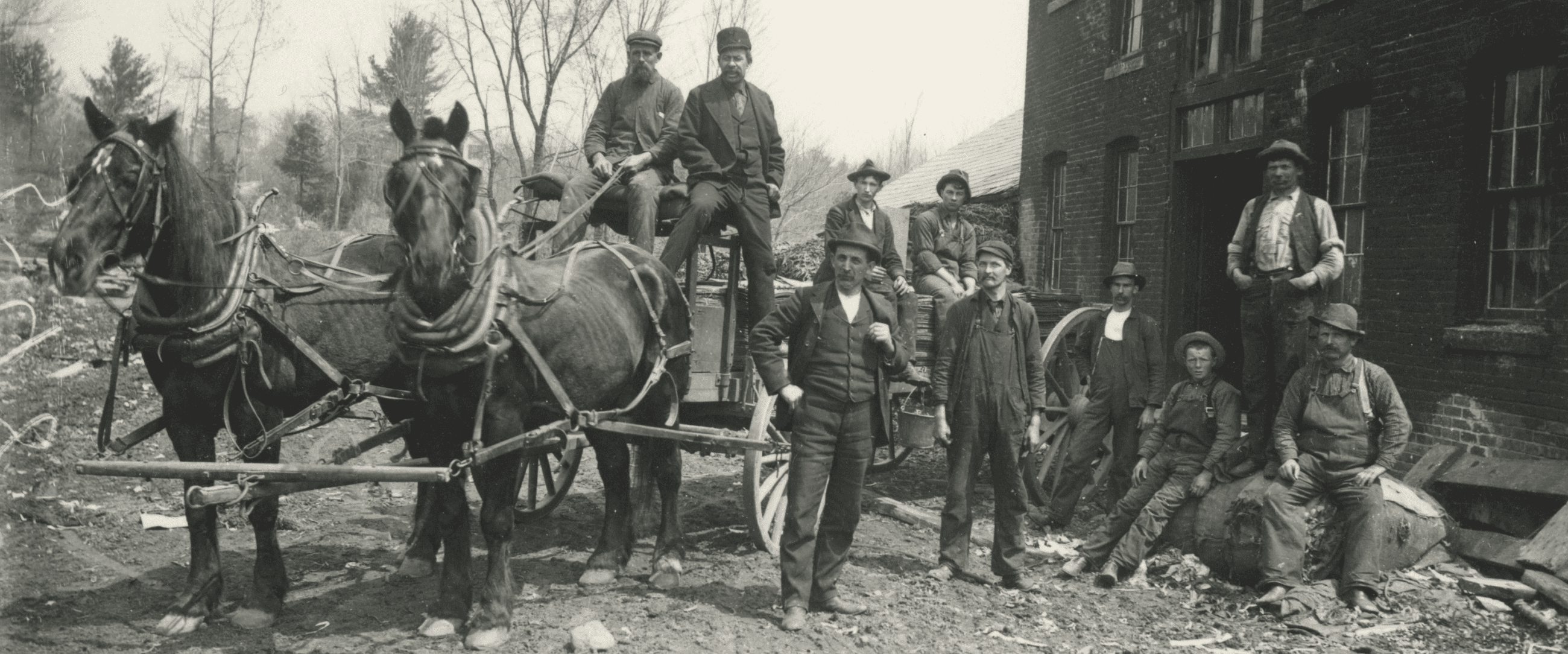 Black-and-white photograph shows eleven men posing with a horse-drawn wagon outside a multi-story brick building. On the left, two dark-colored horses stand harnessed to a wooden wagon, facing left. Two men sit on the elevated driver’s seat of the wagon; the man on the left wears a cap and a knit sweater, while the man on the right wears a brimless cap and a dark suit jacket. Behind them, two more men are seated further back in the wagon bed. To the right of the horses, seven men stand or sit scattered across a dirt ground littered with debris. A man in a dark vest and trousers stands prominently in the foreground near the wagon wheel. Next to him, another man in dark overalls and a cap looks toward the camera. Further right, a younger man sits on a large, wrapped bundle on the ground next to a seated man wearing a brimmed hat. Two other men stand nearby, one leaning against the brick wall of the building. The brick building on the right features multiple tall, narrow windows and an open doorway.