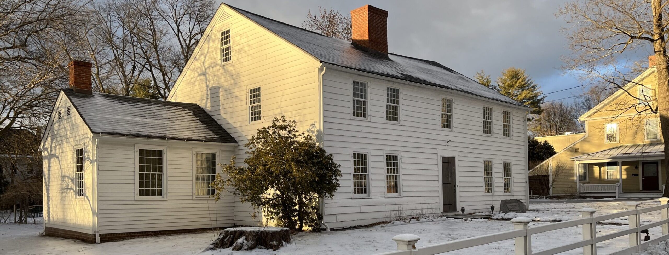 Photograph of a large, two-story historic New England Colonial house featuring white clapboard siding and a central brick chimney. A light snow covers the ground, with a bare tree trunk and small shrubbery positioned directly in front of the house. A low white wooden fence borders the property in the foreground, and another yellow historic house is visible in the background to the right