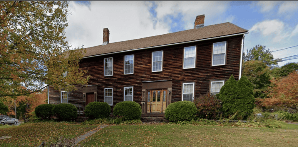 A photograph showing the front facade of a historic two-story house with dark brown wood siding under a cloudy blue sky. The rectangular building features a gabled roof topped with two brick chimneys, flanked by autumn trees with yellow and orange leaves on both sides. A line of five rectangular windows with white frames runs across the second floor, while the first floor features four matching windows alongside a central double wooden door and a smaller red door further to the left. Lush green, rounded bushes line the foundation, and a grassy front yard slopes downward toward the bottom left, where a small section of a paved street and a silver car are visible.