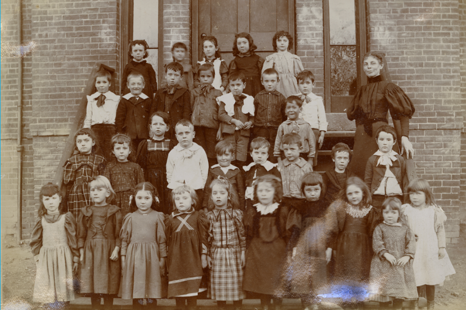A black-and-white photograph shows a large group of young schoolchildren and their female teacher posed on the brick steps of a schoolhouse building. The children, approximately thirty in number and appearing to be between five and ten years old, are arranged in four tiered rows that fill the frame. They wear late 19th- or early 20th-century attire; the girls are dressed in long, dark, often plaid or pinafore-style dresses, while the boys wear dark jackets, button-down shirts with oversized white collars, and trousers. On the far right, a woman stands tall in a dark, high-collared dress with voluminous puffed sleeves, her hair pinned up as she looks toward the camera. The background consists of a textured brick wall with a large wooden door and tall, narrow windows directly behind the group.
