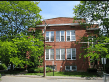 Photograph of a multi-story red brick school building viewed from across a paved street. The symmetrical building features a flat roofline with a raised central parapet and two primary floors over a partially exposed garden-level basement. Each of the main floors displays a wide, central bank of six multi-paned white-trimmed windows grouped in pairs of two, while smaller rectangular windows peek out from the green lawn line at the basement level. In front of the building's left side, a weathered wooden telephone pole stands tall, trailing several black utility lines horizontally across the mid-section of the frame. The structure is flanked and partially shaded by large, mature green trees with vibrant summer leaves, and a low green hedge line runs along the base of the foundation. A small portion of a gray chain-link fence is visible in the lower right corner.