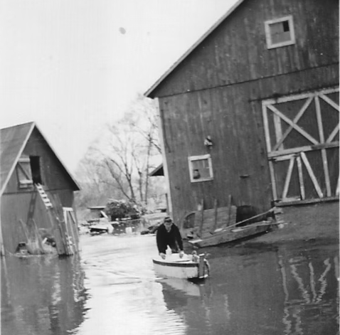 Black-and-white photograph of a flood scene around a pair of rustic wooden barns, with a person navigating the deep waters in a small boat. The right side of the frame is dominated by a large, two-story wooden barn with vertical siding, featuring a small square window on each floor and a large sliding door with a distinct "X" wood brace pattern. Leaning against the base of this barn are loose wooden planks and structural debris, partially submerged by the flood. In the center foreground, a person wearing a dark jacket leans forward inside a small, light-colored motorized boat, steering through the calm, reflective floodwaters that completely submerge the ground. To the left, a smaller wooden shed or barn stands deep in the water, tilting slightly, with a wooden ladder propped up against an open loft door on its side. In the flooded background, bare winter trees and floating debris are scattered across the submerged landscape.