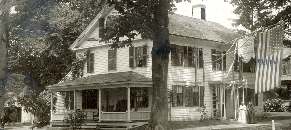 Black-and-white photograph of the front exterior of a large, two-story white wooden house decorated for a historical celebration or civic event. The house features a covered porch on the left side with rocking chairs inside, horizontal clapboard siding, a shingled gabled roof with a central chimney, and numerous multi-paned windows flanked by dark shutters. On the right side, the house is draped with long decorative banners and ribbons, including a large American flag with vertical stripes hanging prominently from an upper branch of a massive, dark tree that stands in the front yard. Near the right entrance pathway, two women dressed in traditional 19th-century period gowns—one in a light dress and one in a dark dress—stand side-by-side facing forward. Lush trees and dense summer foliage frame the entire property.