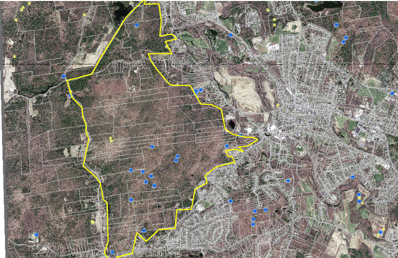 A satillite map displays a large, wooded area outlined by a prominent yellow boundary line. The enclosed territory consists primarily of dense brown and green forest canopy, crisscrossed by straight, narrow property lines or access roads. Scattered throughout this forested zone are several blue circular icons. The yellow border forms an irregular polygon, narrowing significantly at its northern tip and featuring a jagged, pointed indentation on its eastern edge. To the right of the yellow boundary, the landscape transitions into a densely packed, light gray urban development with a tightly woven grid of residential streets, buildings, and houses. Additional blue and yellow icons are scattered sparsely across both the urban neighborhood and the surrounding wooded areas outside the main yellow outline.