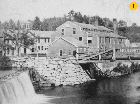 This black-and-white photograph shows a multi-story industrial complex situated along a river. In the foreground, water cascades over a stone-masonry dam on the left, feeding into a river that flows towards the right. A rustic wooden bridge with supporting framework spans across the river from a stone embankment. Several small figures of people stand near the bridge and smaller foreground buildings. The main structures include a prominent two-and-a-half-story gabled wooden mill building with numerous small-paned windows, connected to a long, low-profile industrial wing extending to the right with a row of chimneys. In the background on the far left, another multi-story building features a small cupola or bell tower on its roof. The entire site is framed by a dense backdrop of trees and a rising hillside.
