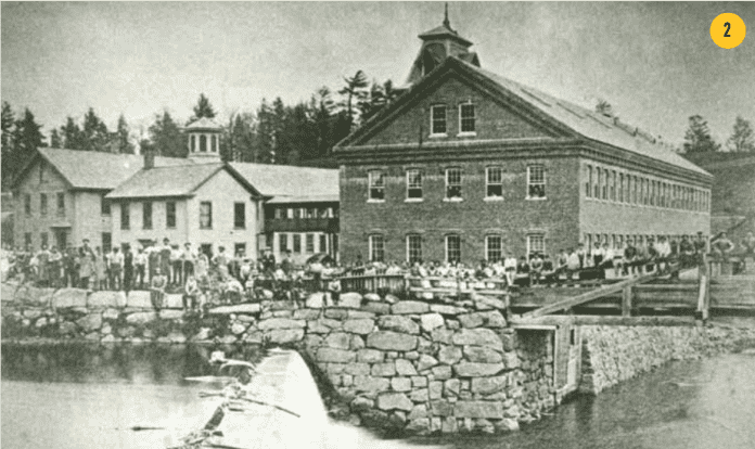 A black and white photograph of an industrial complex situated along a river. On the right, a large two-story brick factory building features a gabled roof topped with a central cupola or bell tower and a long facade lined with rows of rectangular windows. To its left, several smaller connected wooden structures with gabled roofs and individual cupolas extend into the background. A stone retaining wall and a small dam breach the water in the foreground, creating a low waterfall. A large group of workers, appearing as small figures in period clothing, are gathered in a long line across a wooden bridge and along the riverbank in front of the buildings. 
