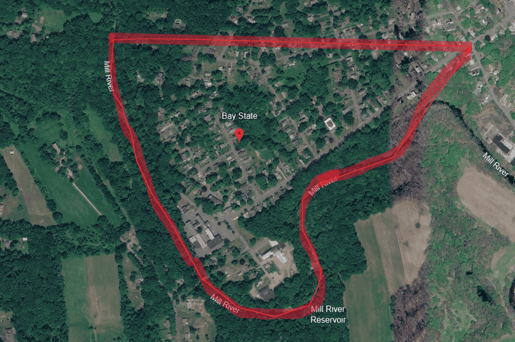 An aerial satellite map views a residential neighborhood outlined by a thick red border. Labeled "Bay State," a red pin marks the center of the neighborhood, which features tree-lined streets, houses, and buildings. The Mill River wraps around the left, bottom, and right borders of the highlighted area, while a straight red line marks the northern boundary. The lower curve of the river is labeled "Mill River Reservoir." Outside the red boundary, dense green forests, open agricultural fields, and neighboring roads are visible.
