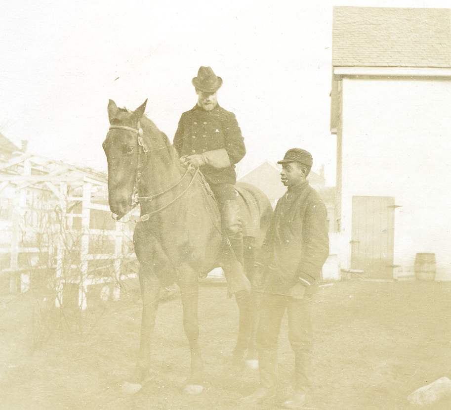 Black-and-white photograph shows a white man riding a horse next to a Black teenage boy standing on the ground outdoors. The man is positioned in the center, mounted on a dark-colored horse that faces left. He wears a dark, double-breasted coat, gloves, high riding boots, and a wide-brimmed hat. He is looking down toward the boy standing to his right. The boy stands on the right side of the frame, facing left toward the rider. He wears a dark jacket, trousers, tall boots, and a structured kepi-style cap. The ground is flat and unpaved. In the background on the left is a white wooden trellis or fence structure. On the right stands a tall, light-colored building with a shingled roof and a closed wooden door.