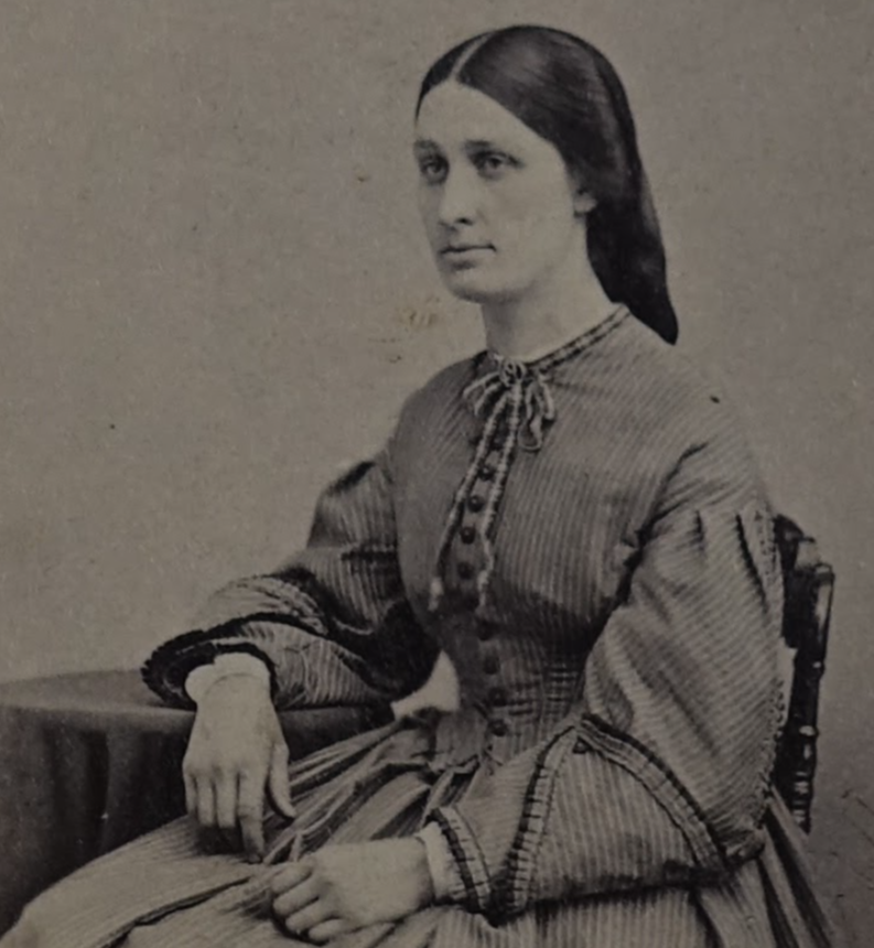 Black-and-white photograph of Sophia Watson. She appears to be in her late 20s or 30s. Her brown hair is styled down. She is wearing a striped dress with puffy sleeves, and leaning one of her arms against a small coffee table.