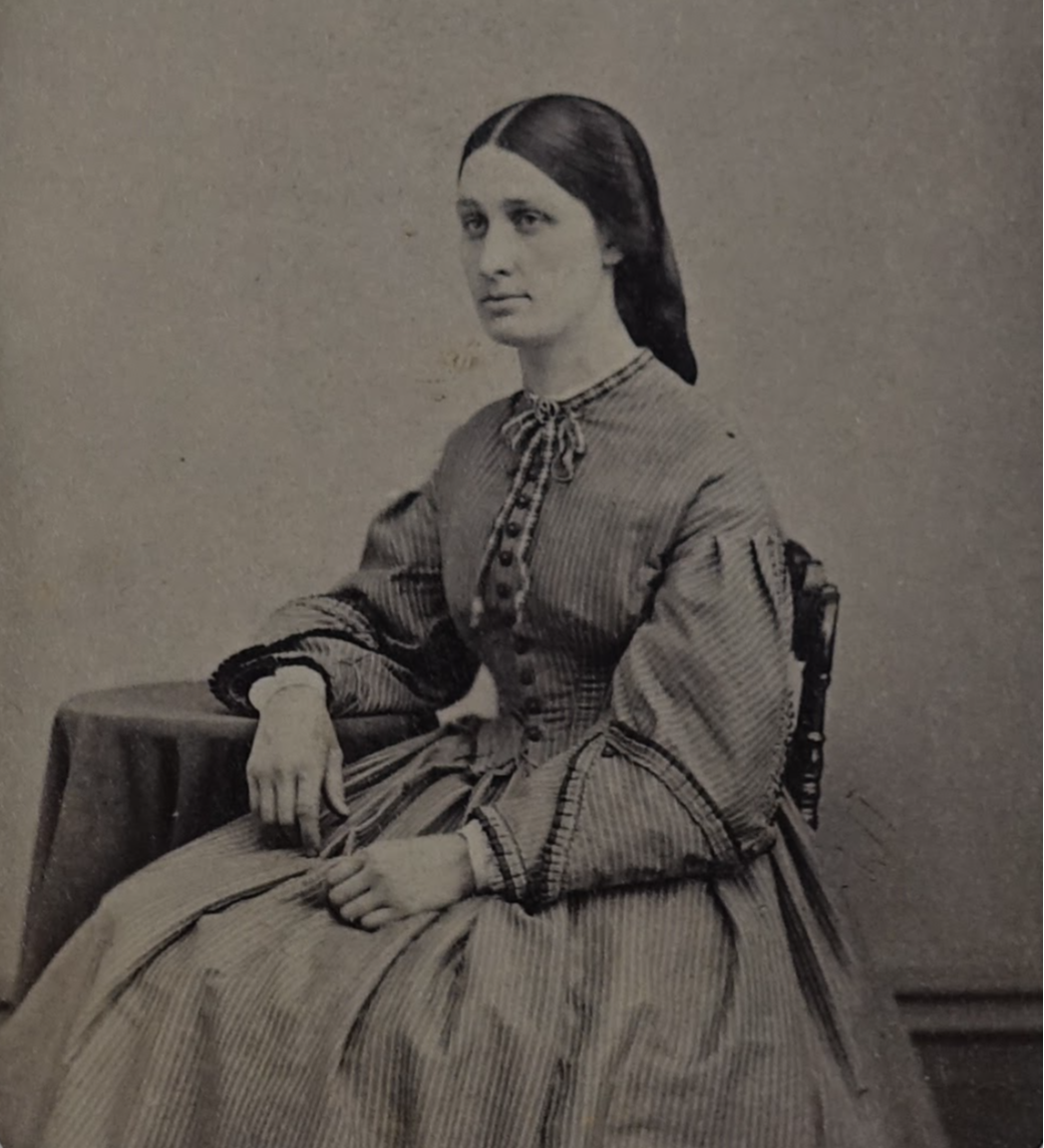 Black-and-white photograph of Sophia Watson. She appears to be in her late 20s or 30s. Her brown hair is styled down. She is wearing a striped dress with puffy sleeves, and leaning one of her arms against a small coffee table.