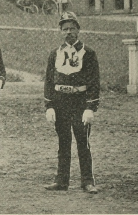 Black-and-white 19th-century photograph of a man with a large mustache in a fireman's uniform, consisting of a helmet; dark shirt with a large collar, white crest with the letter N, and white piping; white gloves; and dark pants with white piping on the side.