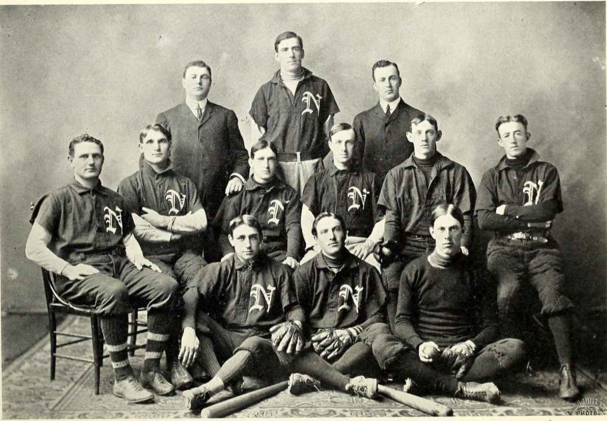 Black-and-white 19th-century photograph of 12 men standing and sitting in three rows, looking forward. Most are wearing baseball uniforms consisting of a short-sleeve collared shirt with a large N on one side of the chest, long sleeves underneath, dark short trousers, and tall dark socks. Two men are wearing dark suits. Three are wearing pitchers gloves. One holds a baseball. There are two baseball bats arranged at the front.