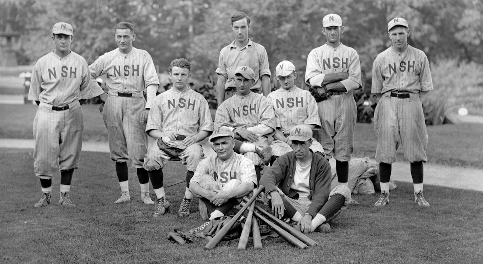 Black and white photograph of a group of 10 men in baseball uniforms with the letters NSH on the front. They are looking forward and posing, some standing and some sitting. In front of them are five baseball bats, criss-crossed over each other and piled on top of a pile of other equipment.