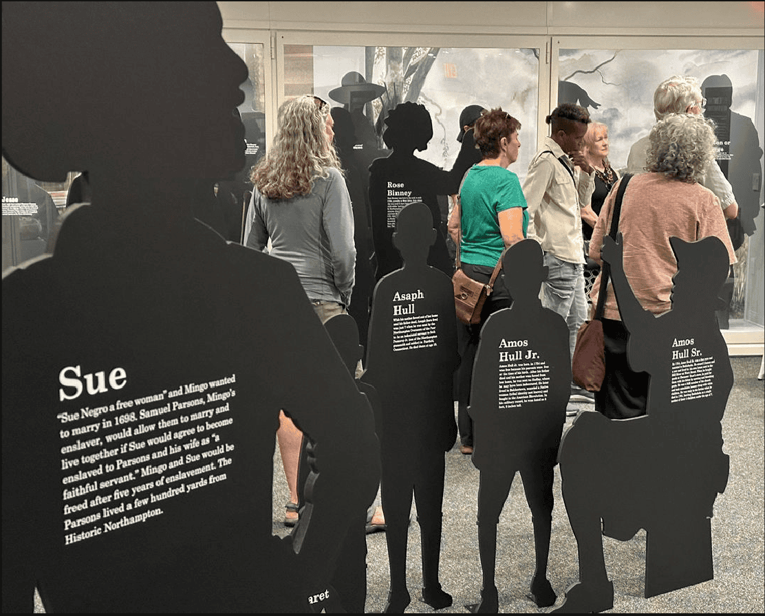 Photograph of museum exhibit with multiple life-sized solid black cut out silhouettes of people. Six people are standing among them, facing away from the camera.
