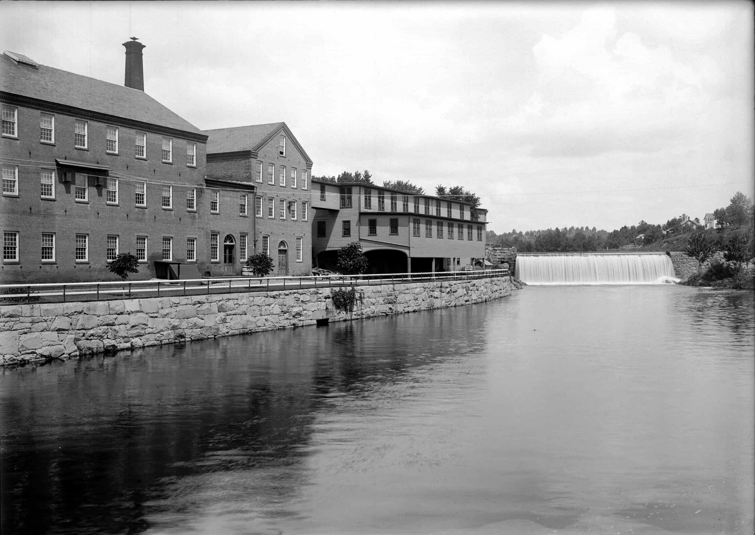 A black and white photograph captures a wide, scenic view of a multi-story industrial brick mill complex situated along the edge of a wide, calm river. The mill buildings feature regular rows of multi-paned windows and are fronted by a low, rustic stone retaining wall lining the riverbank. In the mid-ground, a wide dam stretches across the river, creating a smooth, white cascade of water that spills into the lower section of the river.  The far bank in the distance is lined with rolling hills and clusters of trees.