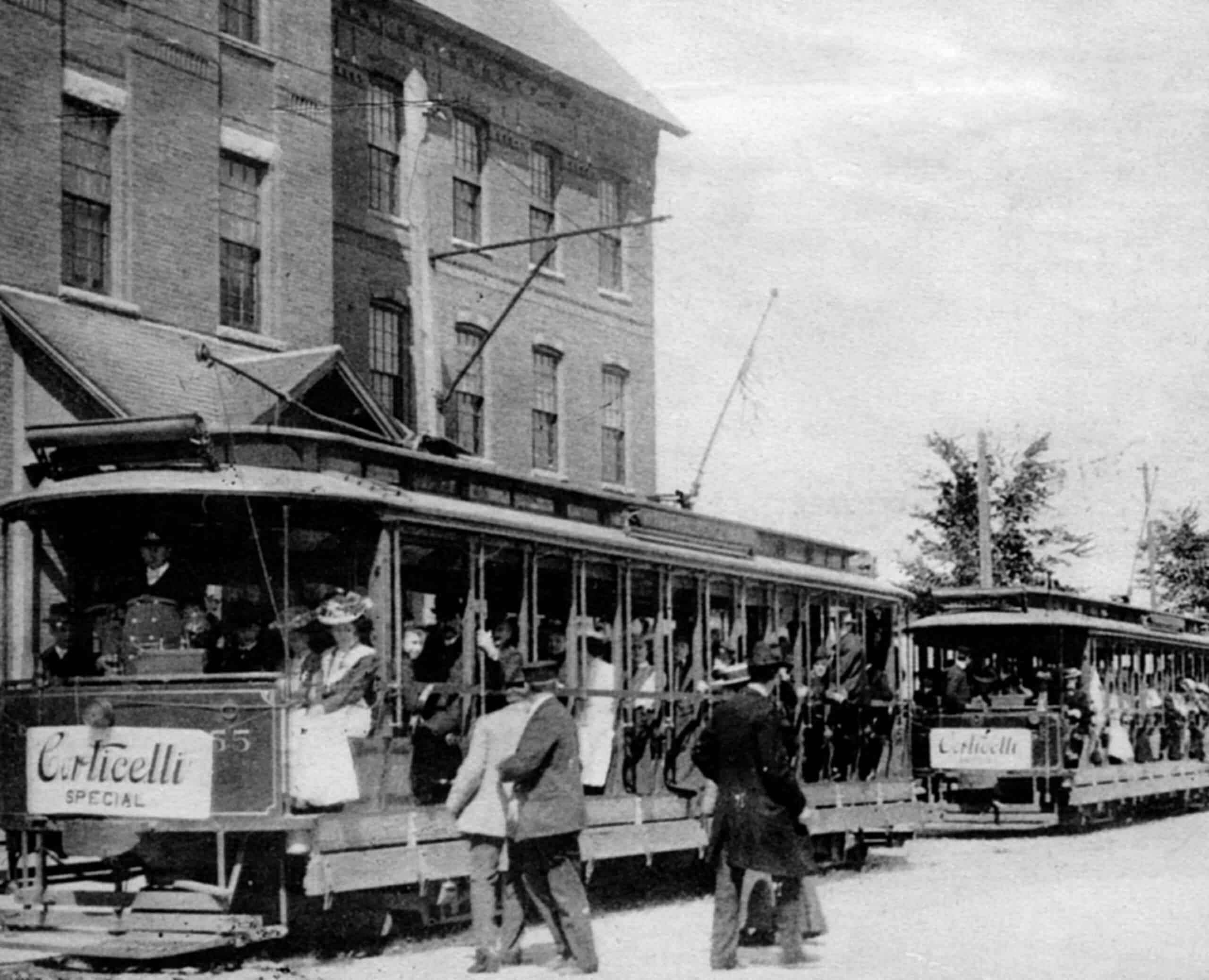 A black and white photograph shows two open-sided, electric streetcars packed with passengers dressed in early 20th-century attire. The streetcars are stopped on a dirt street next to a multi-story brick building with rows of rectangular windows. The streetcar in the foreground has a prominent white sign on its front bumper that reads "Corticelli SPECIAL" in a cursive script, with a matching sign visible on the front of the second streetcar further back. Passengers, including women in wide-brimmed hats and long skirts, are seated inside the open compartments, while several men in suits and hats stand on the street beside the streetcars. Overhead trolley poles extend from the roofs of the streetcars to connect with electrical wires running along the side of the building.