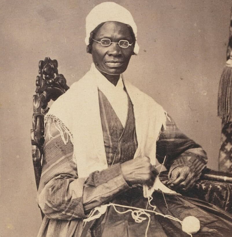 Black-and-white photographic portrait of Sojourner Truth sitting in a chair. She has a ball of yarn on her lap and is knitting. She wears a dark dress with a white material draped on her shoulders and she has a white bonnet or cap on her head.