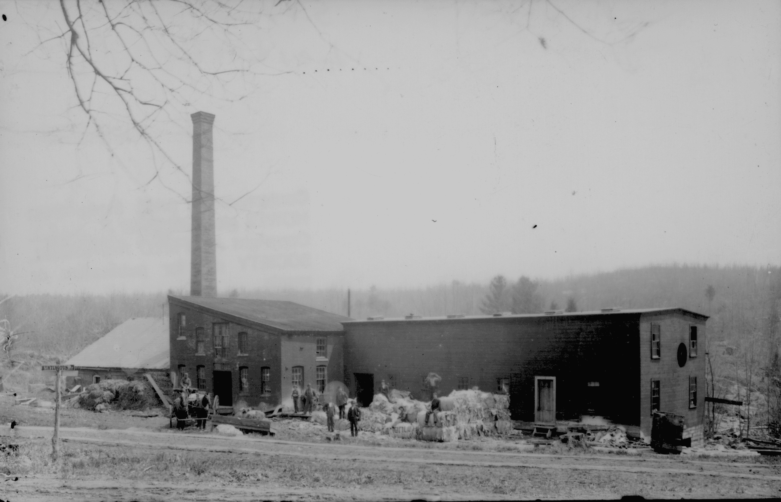 A black-and-white photograph captures an old industrial facility, against a rural landscape. The multi-part complex features a prominent, tall brick smokestack rising from the rear of a two-story brick building on the left. This central building has a slanted roofline, multiple multi-pane windows, and open doorways. To its left, a low, single-story structure with a light-colored, angled roof extends outward. On the right side of the complex stands a long, two-story wooden building with dark siding and sparse window placement. In front of the buildings, several individuals are scattered along a dirt path or clearing. A small horse-drawn cart is positioned near the left building entrance. Large, bundled rectangular stacks are piled on the ground outside the wooden structure. The foreground consists of an open dirt road, while the background reveals a dense, rolling treeline. Sparse, bare tree branches hang into the upper-left corner of the frame.