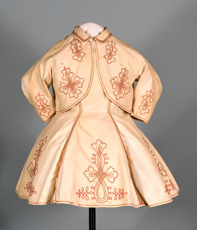 Child's dress likely for a boy made of white pique cotton with a matching jacket, circa 1866-1870. The dress is constructed with modified princess seams from the armscye to the hem in front and back. The seams in front are outlined with pink and tan soutache braid (the pink was originally red). The short sleeves are ruffled with pleats in the armscye. The opening edge is scalloped at the shoulder. The neckline is wide and round. The sleeves and the skirt are decorated with arabesque medallions of soutache. The center back opening extends 11.5" and fastens with five vegetable ivory ball buttons on metal shanks. The jacket has a cutaway front and it fastens with one white china shank button and cord loop at the base of the neck under the turned down collar. The sleeves are cut as a coat sleeve. The jacket fronts sleeves and lower center back are embellished with arabesque medallions of soutache.