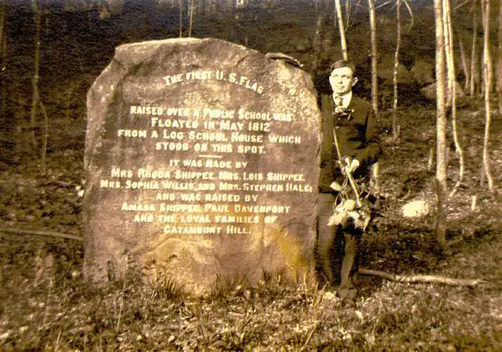 A black-and-white photograph of a large, upright stone monument set against a wooded background. Standing to the right of the stone is a young man in a dark suit and tie, holding a small bundle of plants or flowers. The stone features an engraved historical inscription that reads:"THE FIRST U.S. FLAG RAISED OVER A PUBLIC SCHOOL WAS FLOATED IN MAY 1812 FROM A LOG SCHOOL HOUSE WHICH STOOD ON THIS SPOT. IT WAS MADE BY MRS. RHODA SHIPPEE, MRS. LOIS SHIPPEE, MRS. SOPHIA WILLIS AND MRS. STEPHEN HALE, AND WAS RAISED BY AMASA SHIPPEE, PAUL DAVENPORT AND THE LOYAL FAMILIES OF CATAMOUNT HILL."