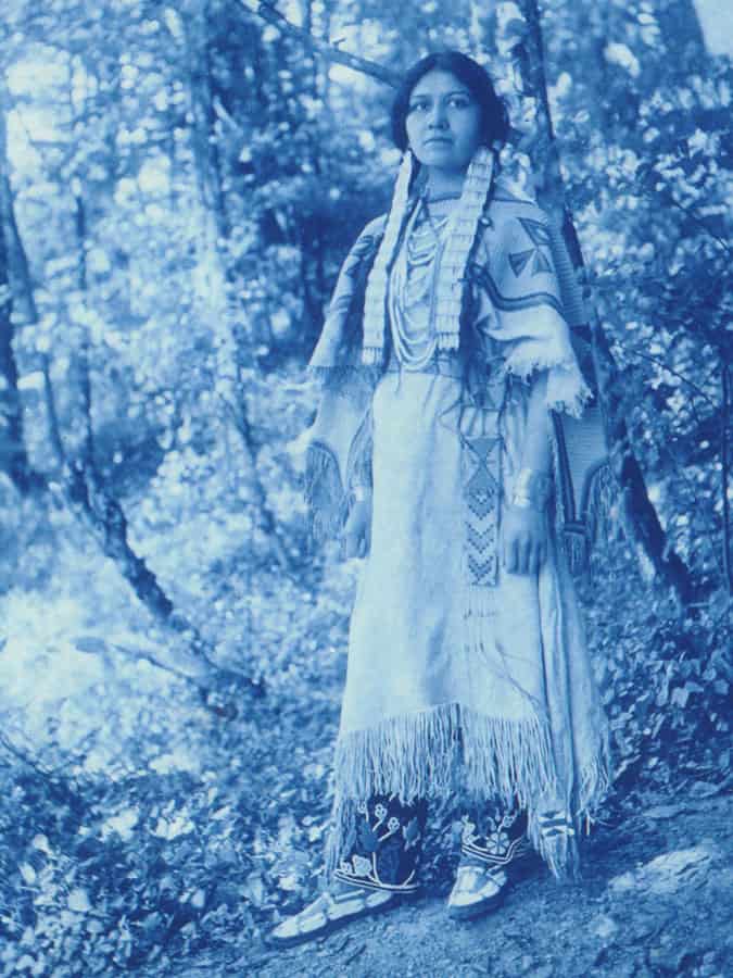 Blue-tinted photograph (cyanotype) of a young woman in traditional Native American dress stands on a hill in sun-dappled woods.