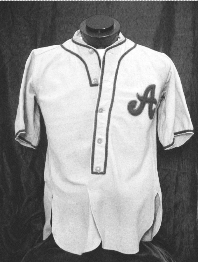 Black-and-white photograph of an old-fashioned baseball jersey. It is white with dark piping around the button-front, neckline, and sleeve. The is a cursive A on one side of the chest.