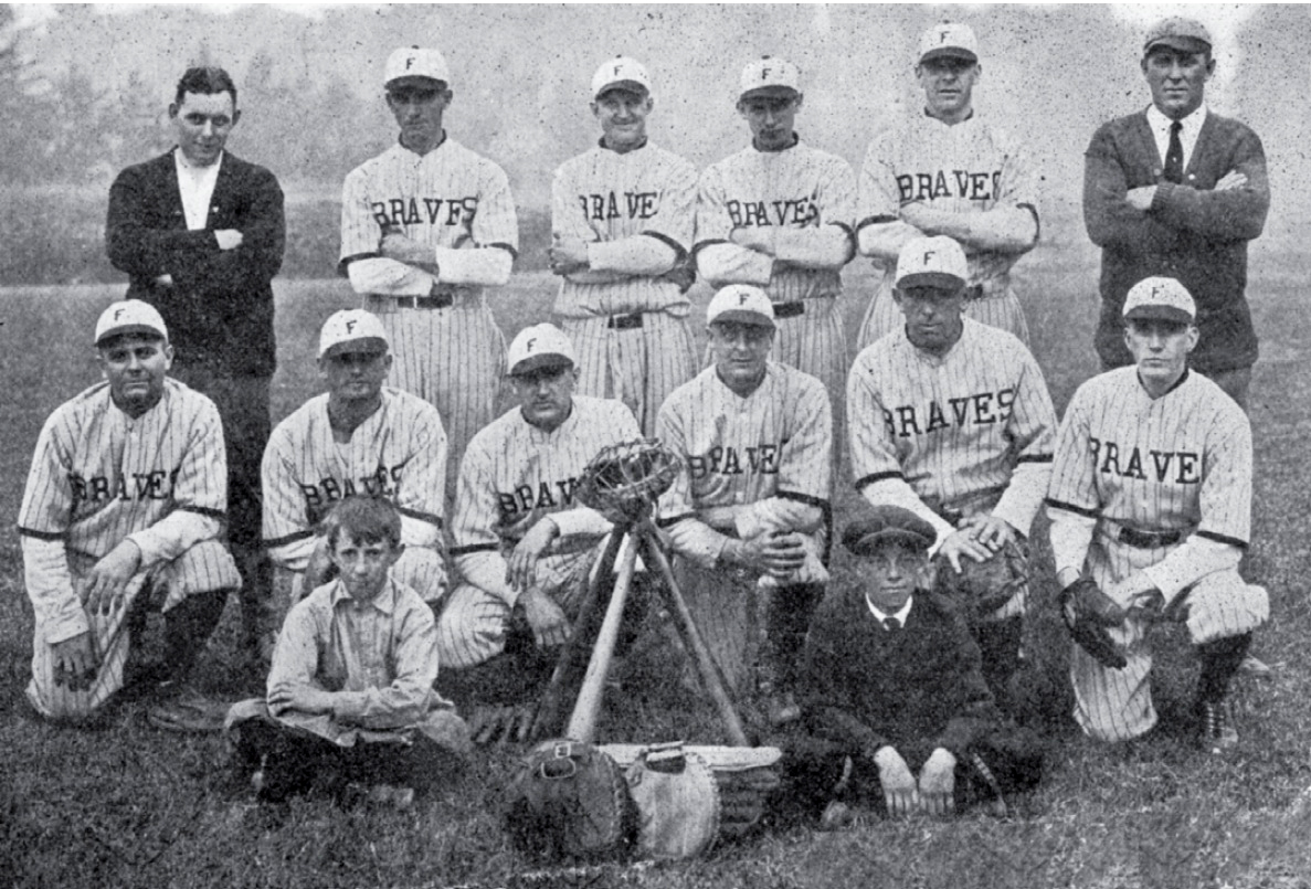 Black-and-white early 20th-century photograph for 12 men and two young boys standing with arms crossed or kneeling in two rows, with the two boys seated to the front. Two men are wearing cardigans and the rest are wearing baseball uniforms consisting of white hats with dark brims as the letter F in them, pinstripe light shirts with "Braves" on the front, and striped pants. Bats and other equipment are arranged in the front.